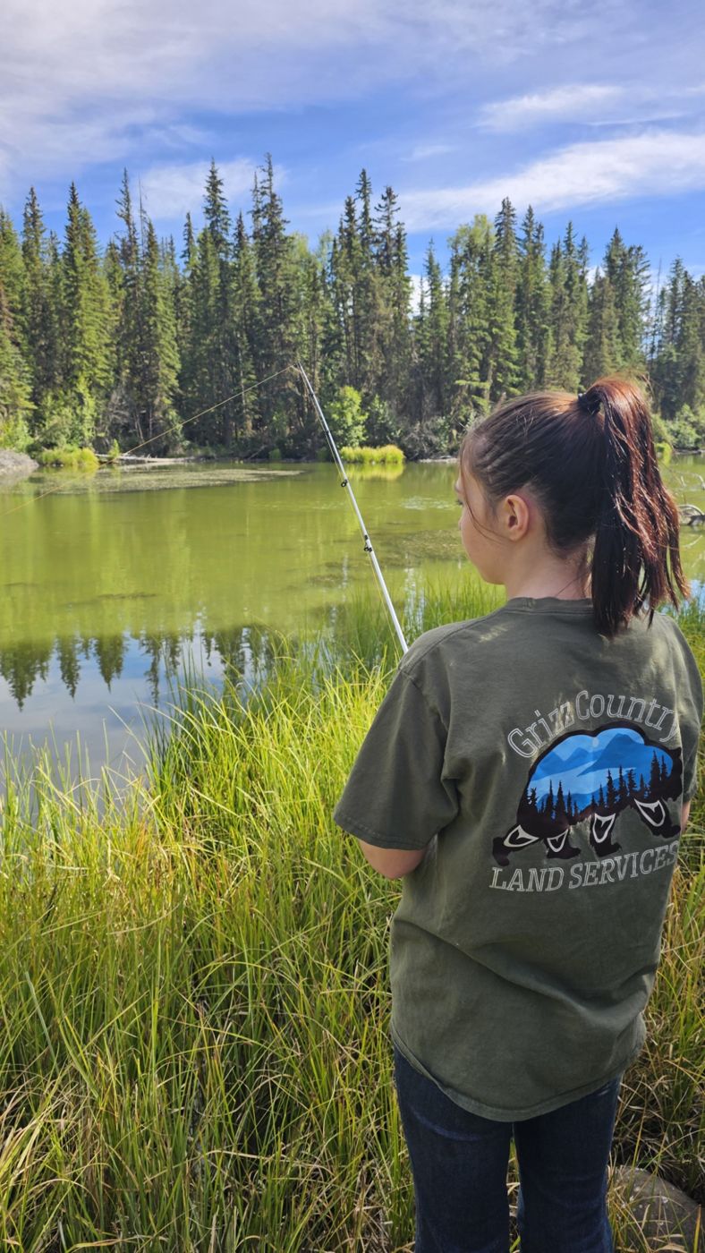 A kid in a Grizz Country shirt fishing a green lake edged with spruce